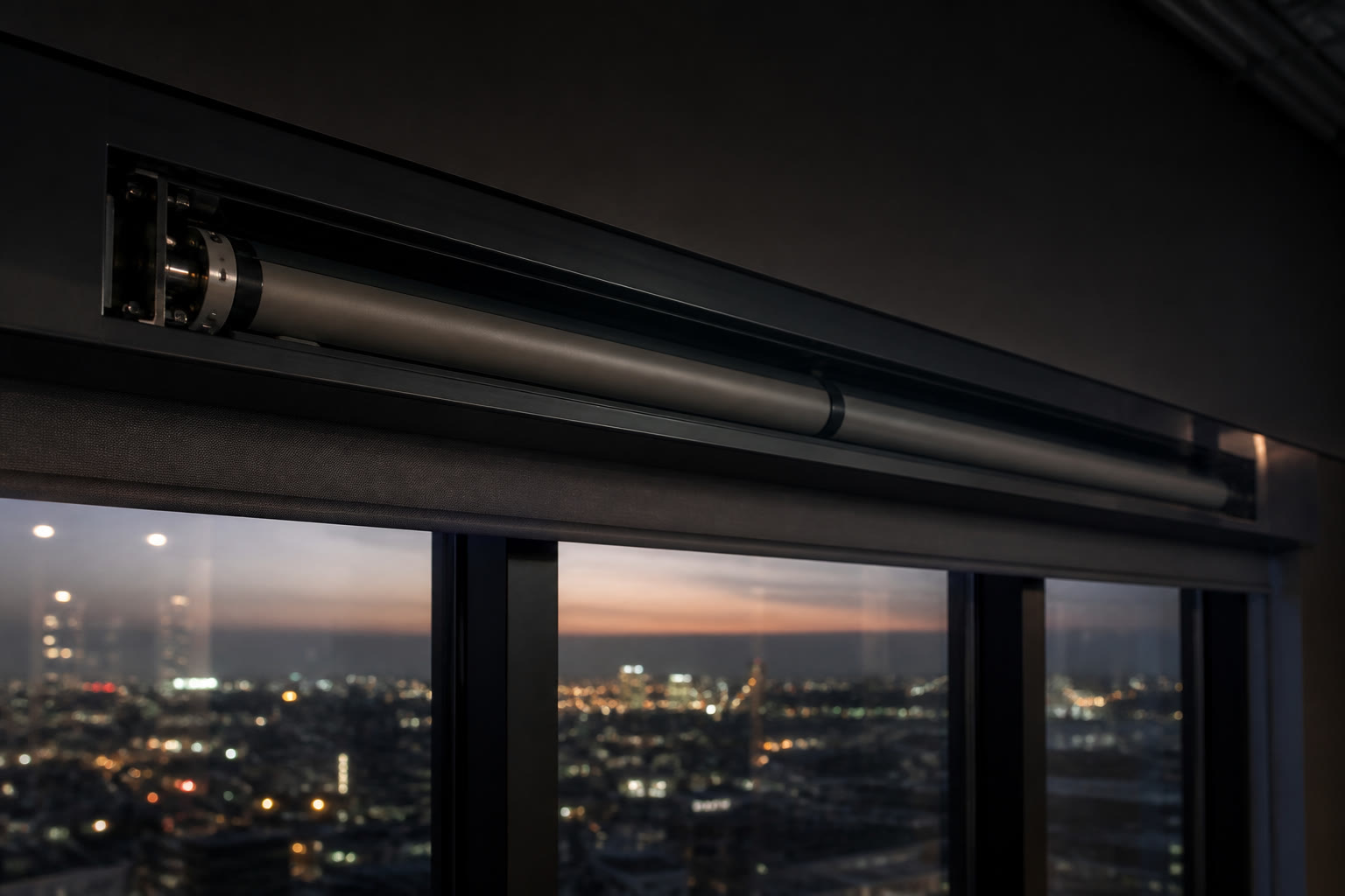 The head rail of a motorised roller blind at the top of a floor-to-ceiling window at dusk, the tubular motor visible in the pocket with city glow through the glass.