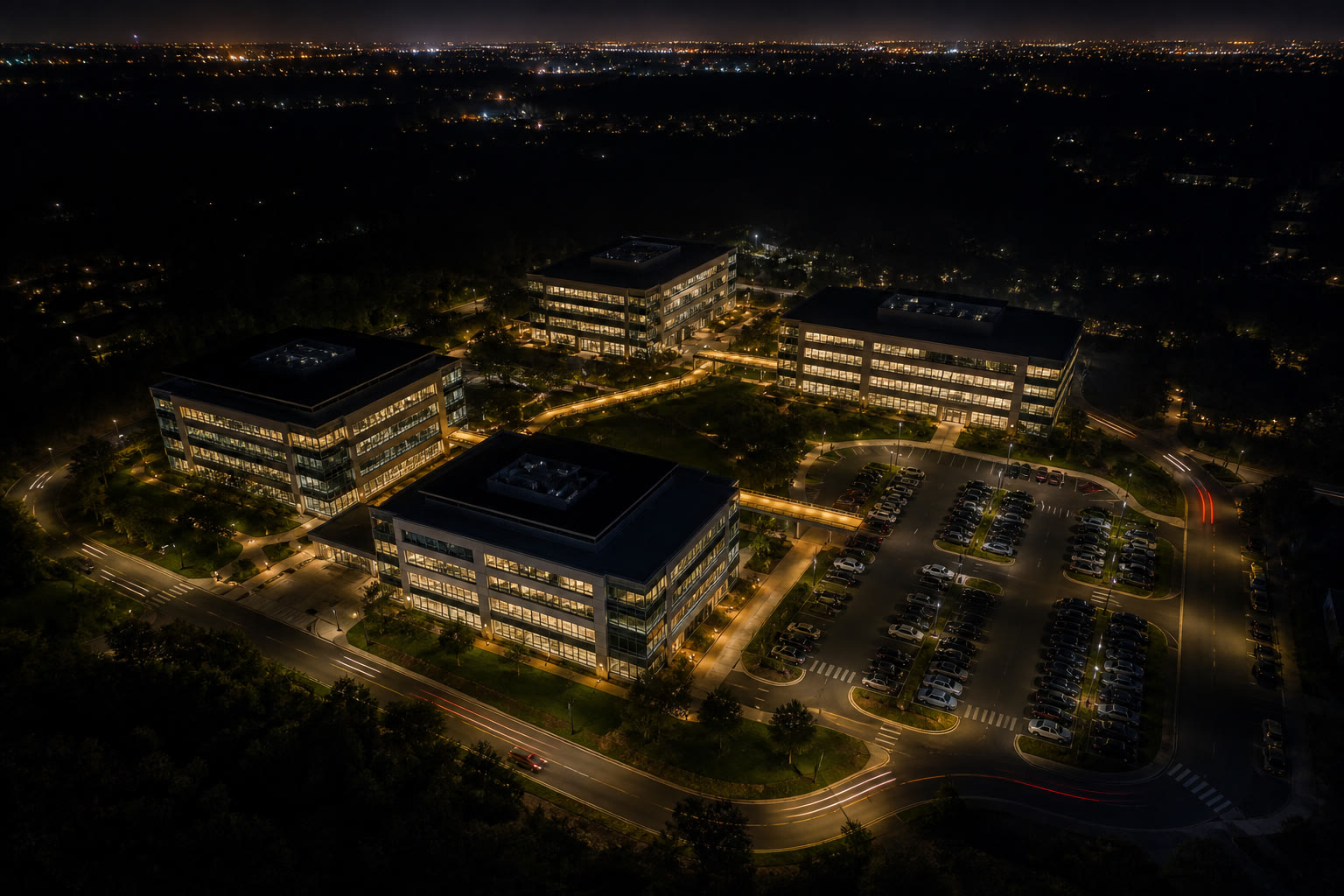 An aerial night view of a corporate campus of several mid-rise buildings linked by lit walkways and a car park, with city glow on the horizon.