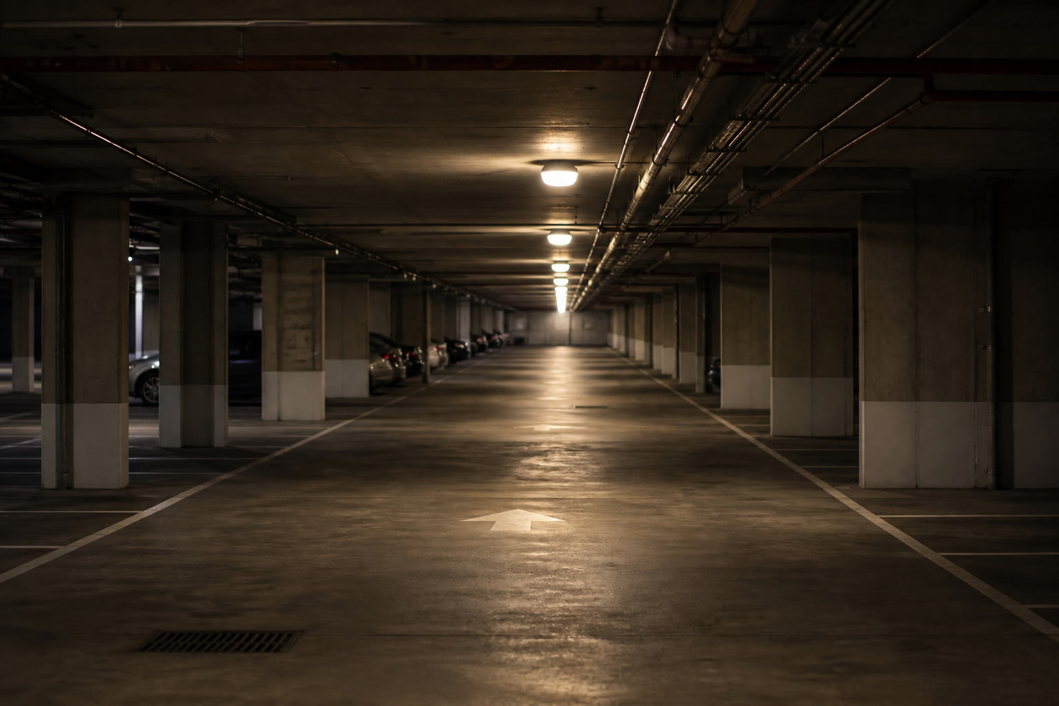 An underground car park level at night, evenly spaced ceiling luminaires receding down the aisle, painted bay markings and exposed services overhead.
