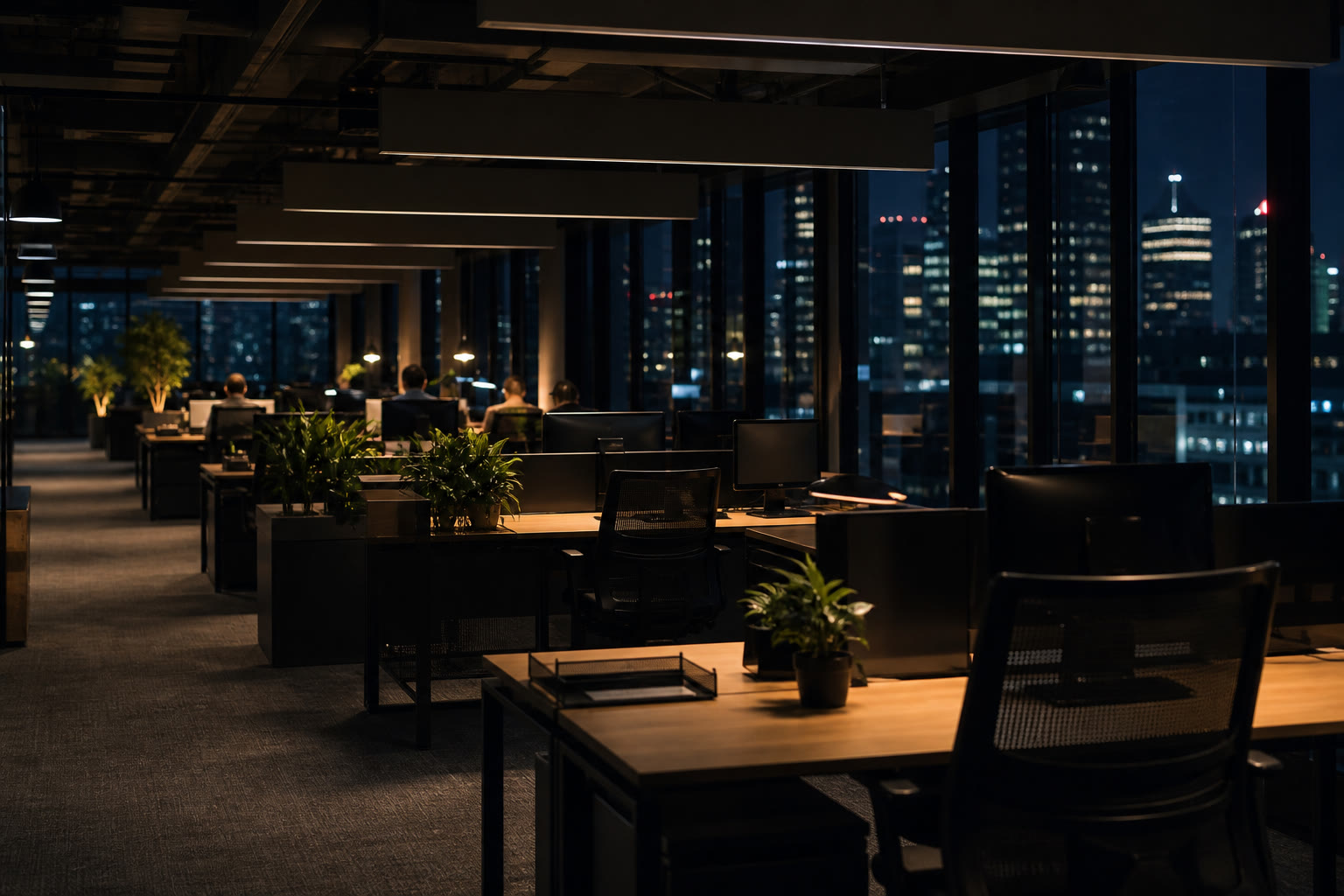 An occupied open-plan office floor at dusk with desks, chairs and monitors turned away, warm even lighting and a dark city beyond the glazing.